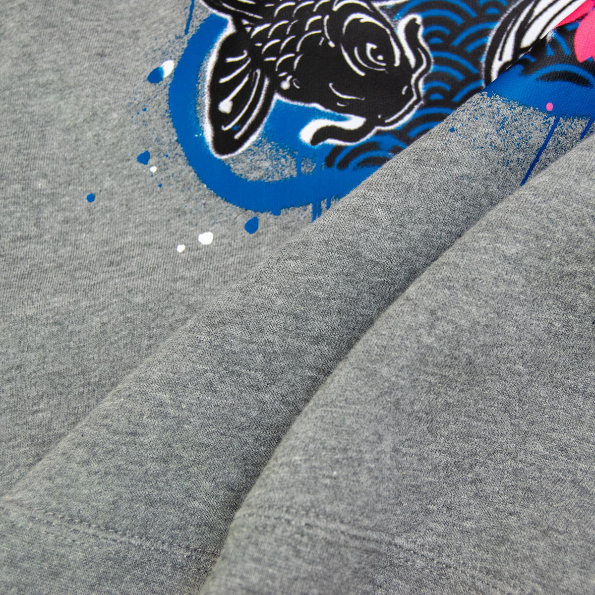 Close-up of a heather gray fabric garment featuring a graphic print of a black koi fish, blue waves, and pink cherry blossoms.
