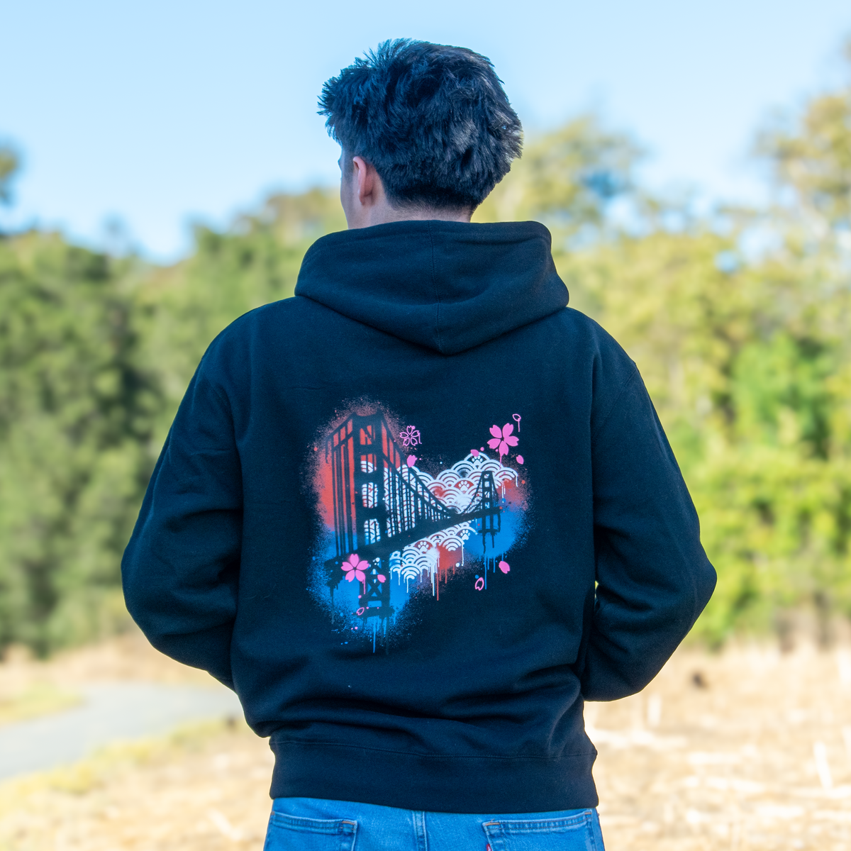 A person viewed from behind wearing a black hoodie with a colorful graphic print of the Golden Gate Bridge and cherry blossoms on the back, standing outdoors in a park with trees and dry grass.