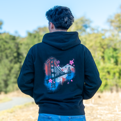 A person viewed from behind wearing a black hoodie with a colorful graphic print of the Golden Gate Bridge and cherry blossoms on the back, standing outdoors in a park with trees and dry grass.