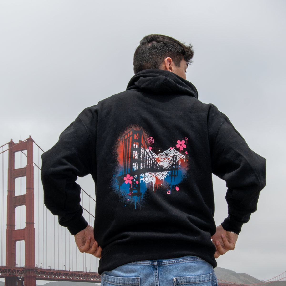 A man viewed from behind wearing a black hoodie with a colorful graphic print of the Golden Gate Bridge on the back, standing in front of the bridge under an overcast sky.