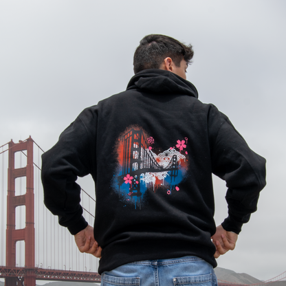 A man viewed from behind wearing a black hoodie with a colorful graphic print of the Golden Gate Bridge on the back, standing in front of the bridge under an overcast sky.
