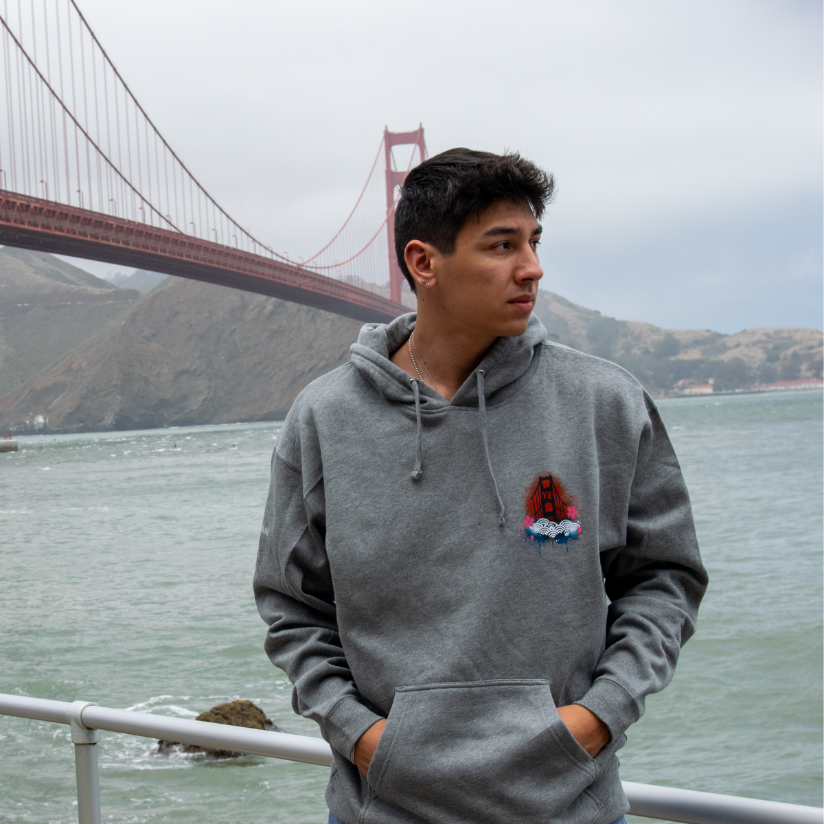 A man wearing a gray pullover hoodie with a small red, white, and blue graphic on the left chest stands outdoors with his hands in his pockets, with the Golden Gate Bridge and water in the background.