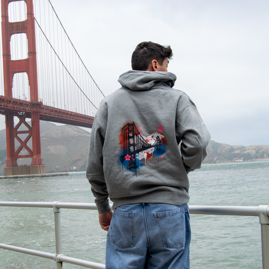 A man viewed from behind wearing a gray hoodie with a colorful graphic print on the back, standing in front of the Golden Gate Bridge over the water.