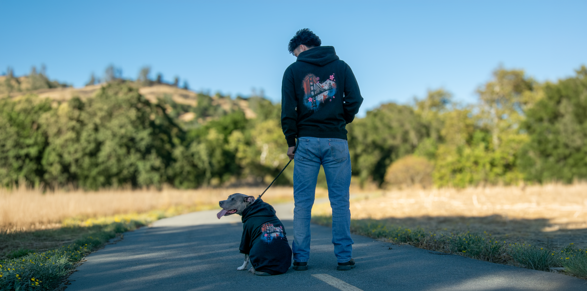 A man and a dog wearing matching black graphic hoodies stand on a paved path outdoors, with trees, grass, and a blue sky in the background.