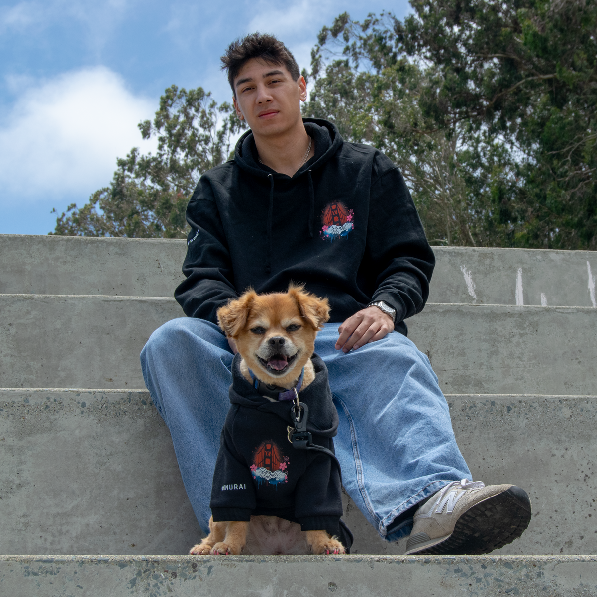A man and his dog sitting on concrete stairs outdoors. Both are wearing matching black hoodies with a red and blue graphic on the chest and white text near the hem.