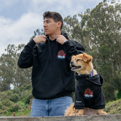A man and a dog wearing matching black hoodies with a red and blue graphic logo, standing outdoors on a wooden picnic table with trees and sky in the background.