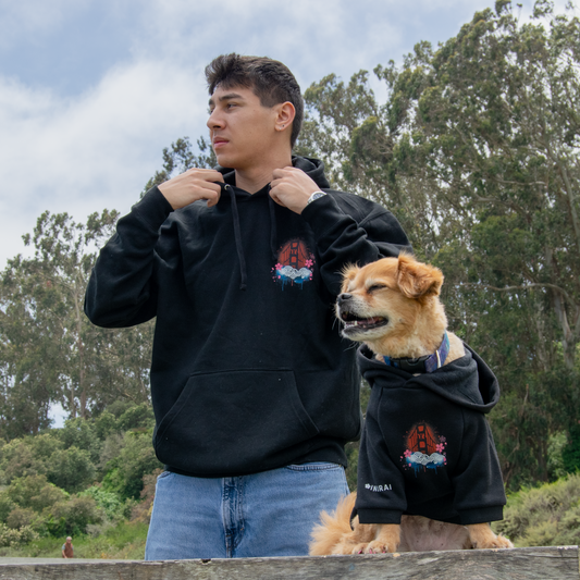 A man and a dog wearing matching black hoodies with a red and blue graphic logo, standing outdoors on a wooden picnic table with trees and sky in the background.