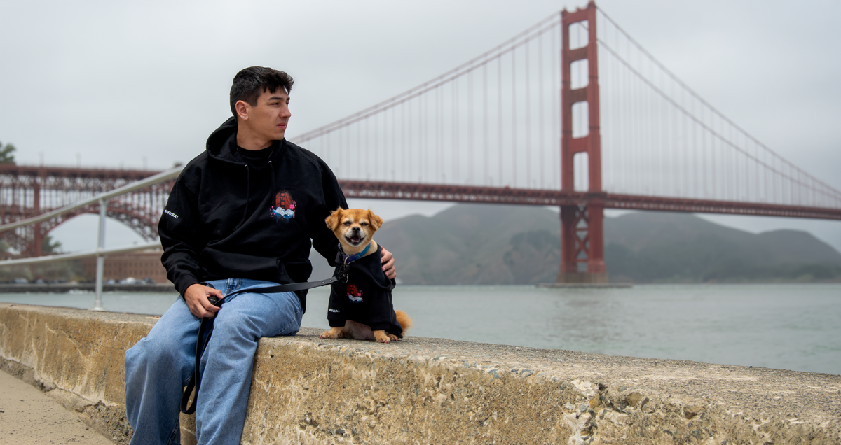 A man wearing a black hoodie and blue jeans sits on a concrete ledge next to a small tan dog wearing a matching black hoodie, with the Golden Gate Bridge and water in the background.