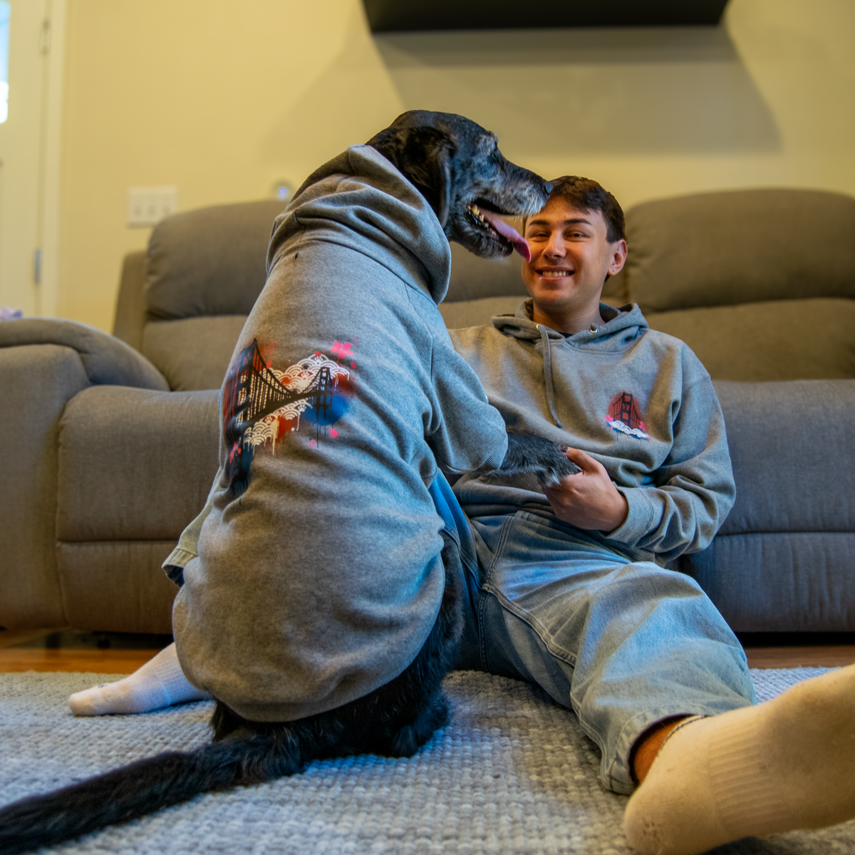 A man in a matching gray hoodie and sweatpants sits on a carpeted floor in a living room, petting a black dog wearing a gray hoodie with a golden gate bridge graphic on the back.