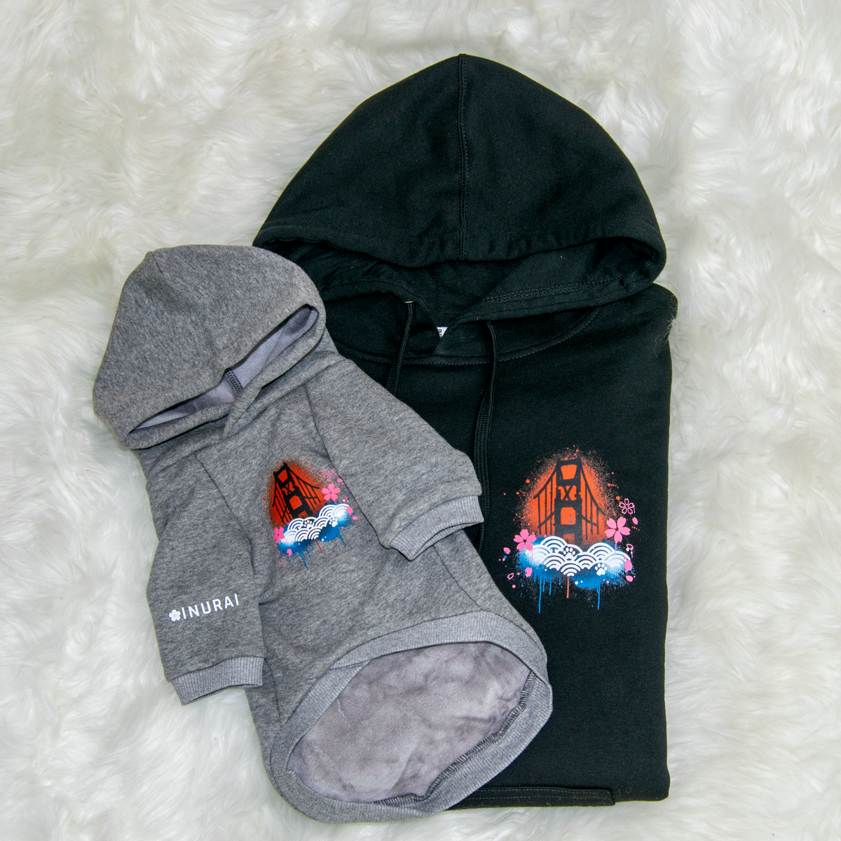 Two hooded pet sweatshirts laid flat on a white faux-fur surface: a black hoodie with a colorful Golden Gate Bridge graphic on the front and a gray hoodie placed on top with a matching bridge graphic and small white text on the sleeve.