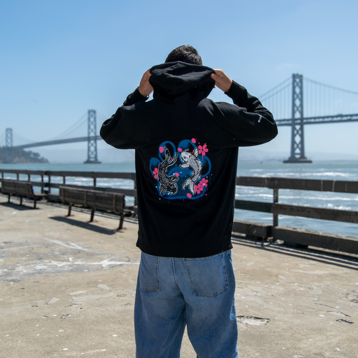 A man stands on a waterfront promenade wearing a black hoodie with a large back graphic of two fish in a blue heart with pink flowers, with the Bay Bridge in the background.