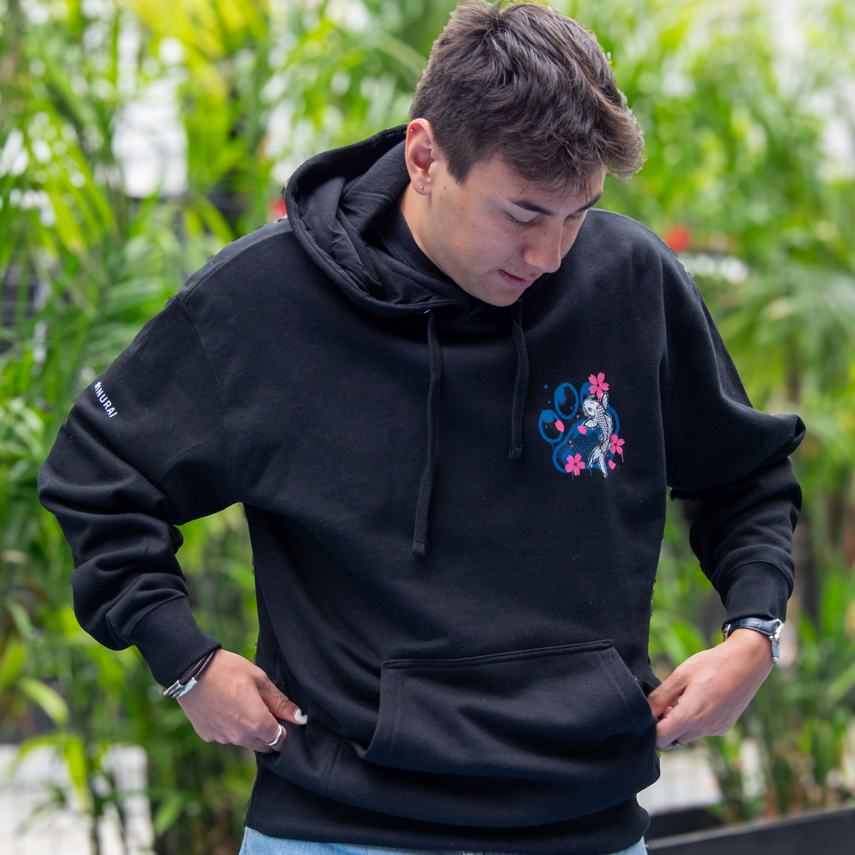 A man wearing a black pullover hoodie with a colorful graphic on the left chest, standing in front of a blurred background of green plants.