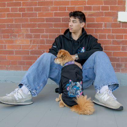 A man and a small brown dog sitting wearing matching black sweatshirts leaning against a brick wall