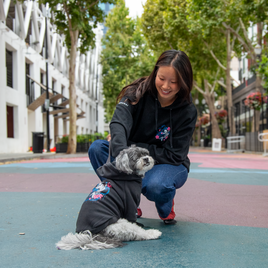 A woman crouches on a colorful outdoor street wearing a black hoodie with a dog on it, also wearing a matching black hoodie and jeans. The dog is wearing a small black hoodie with a colorful graphic.
