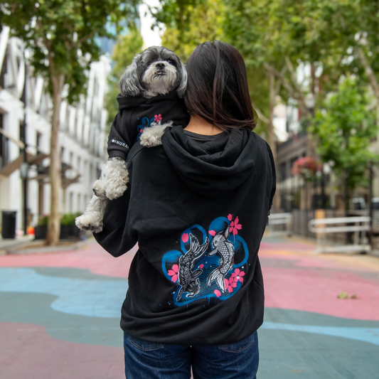 A woman from behind wearing a black hoodie with a large blue and pink koi graphic on the back, holding a small gray dog dressed in a matching black hoodie. They are standing on a colorful paved street in an outdoor urban setting with trees and buildings in the background.