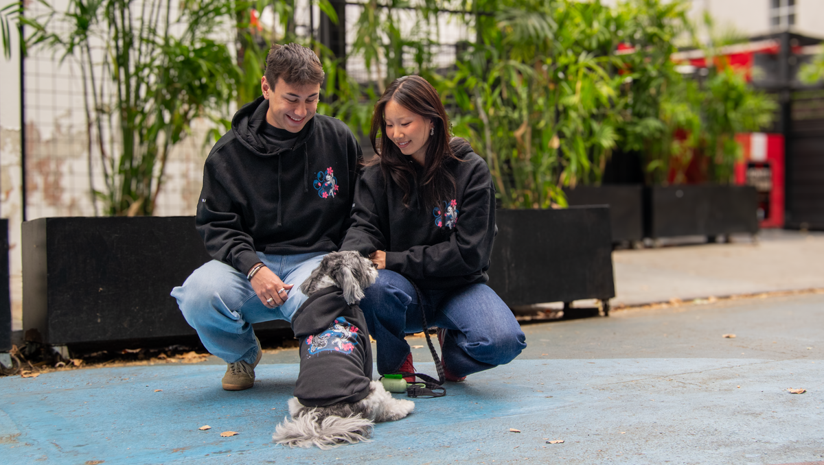 A man and a woman crouching outdoors next to a small gray dog, all wearing matching black hoodies with a colorful graphic on the chest. The scene is set on a blue paved path with potted plants in the background.