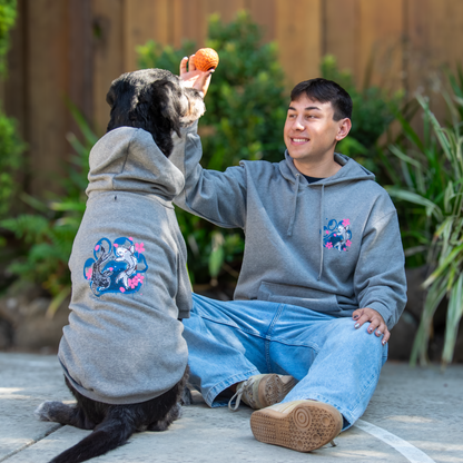 A man and his black dog sit together outdoors wearing matching gray hoodies with colorful floral and tiger graphics on the back. The man offers a treat to the dog, with plants and a wooden fence in the background.