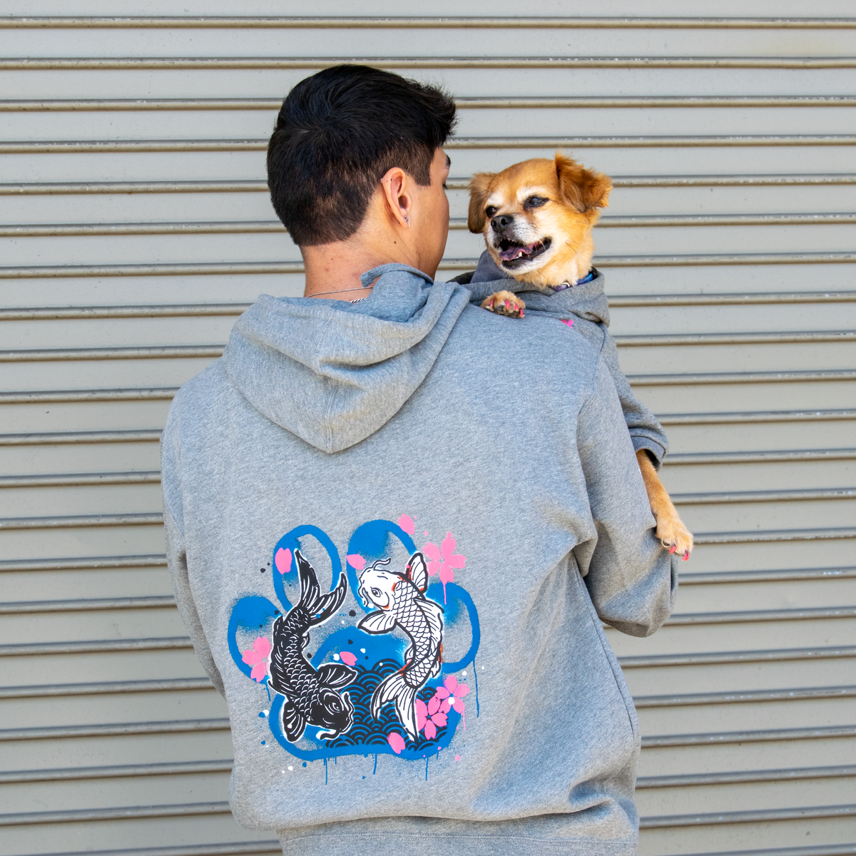 A man wearing a light gray hoodie with a colorful koi fish graphic on the back, holding a small tan dog against a gray metal shutter background.