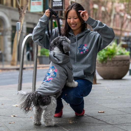 A woman crouches on a city sidewalk next to a small gray and white dog, both wearing matching gray hoodies with a colorful floral graphic on the chest. The background shows a blurred urban street scene with planters and a lamp post.