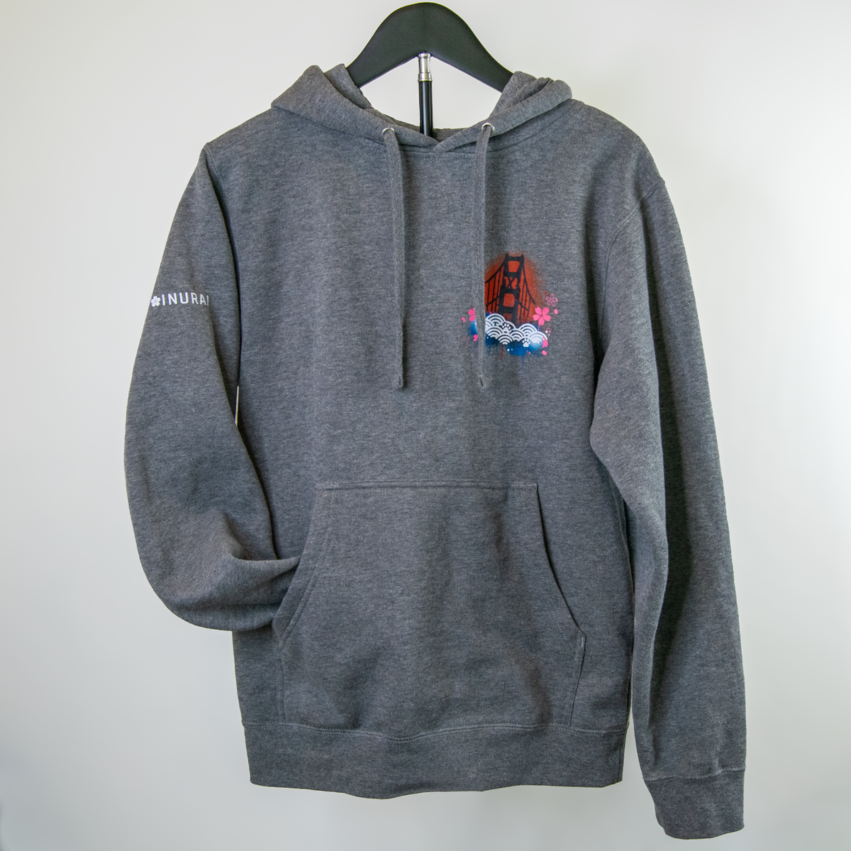 A dark gray heather pullover hoodie hanging on a black hanger against a plain light gray wall. The hoodie features a small multicolor graphic on the left chest depicting the Golden Gate Bridge with flowers