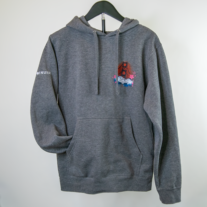 A dark gray heather pullover hoodie hanging on a black hanger against a plain light gray wall. The hoodie features a small multicolor graphic on the left chest depicting the Golden Gate Bridge with flowers