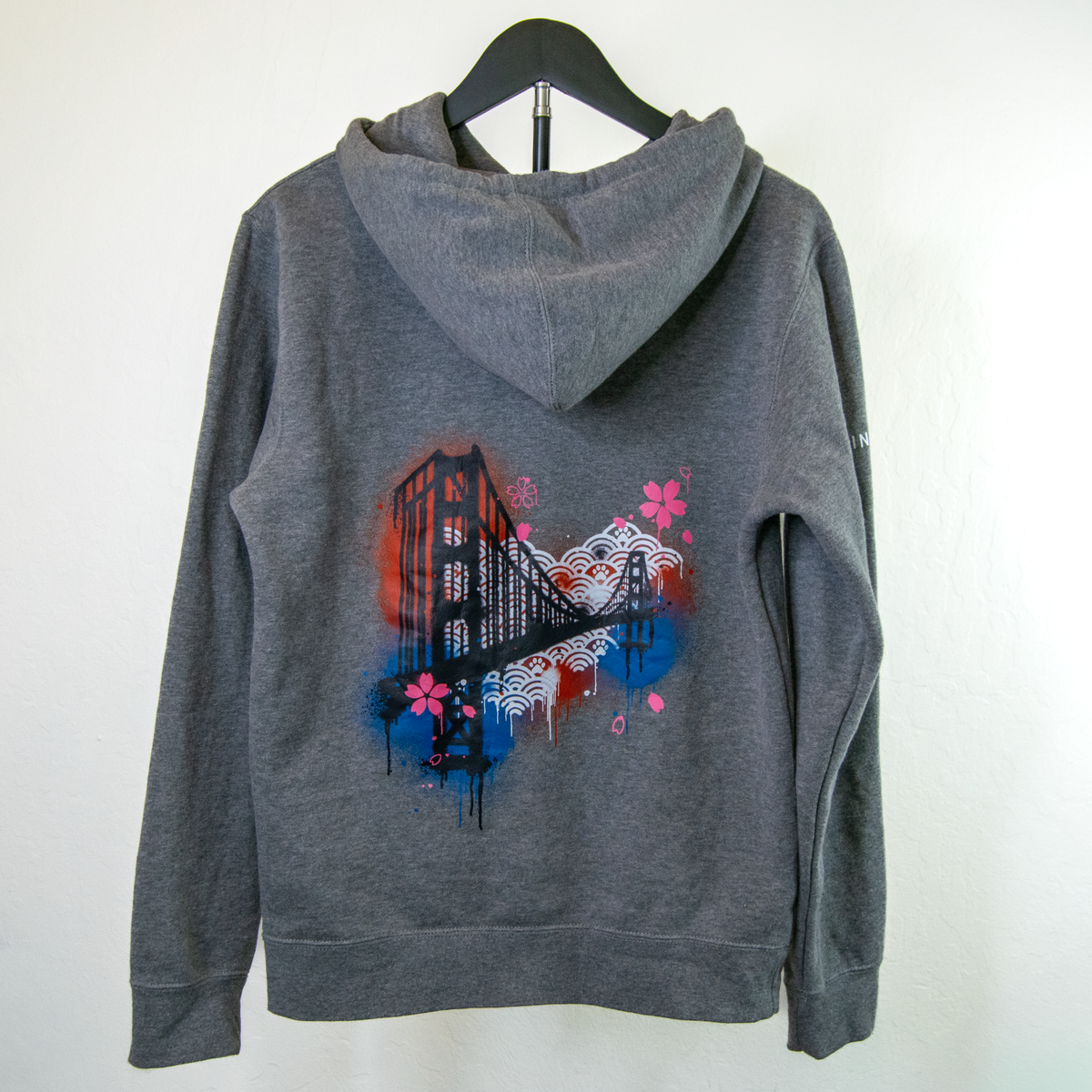 A dark gray heathered hoodie hanging on a black hanger against a white wall, featuring a colorful graphic print on the back with the golden gate bridge, waves, and pink cherry blossoms.