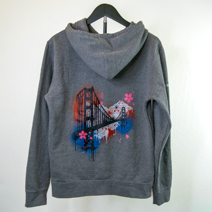 A dark gray heathered hoodie hanging on a black hanger against a white wall, featuring a colorful graphic print on the back with the golden gate bridge, waves, and pink cherry blossoms.