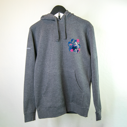 A dark gray heather pullover hoodie hanging on a black hanger against a plain off-white wall. The hoodie features a small colorful graphic on the left chest depicting a koi fish with pink flowers, and a small white logo on the right sleeve.