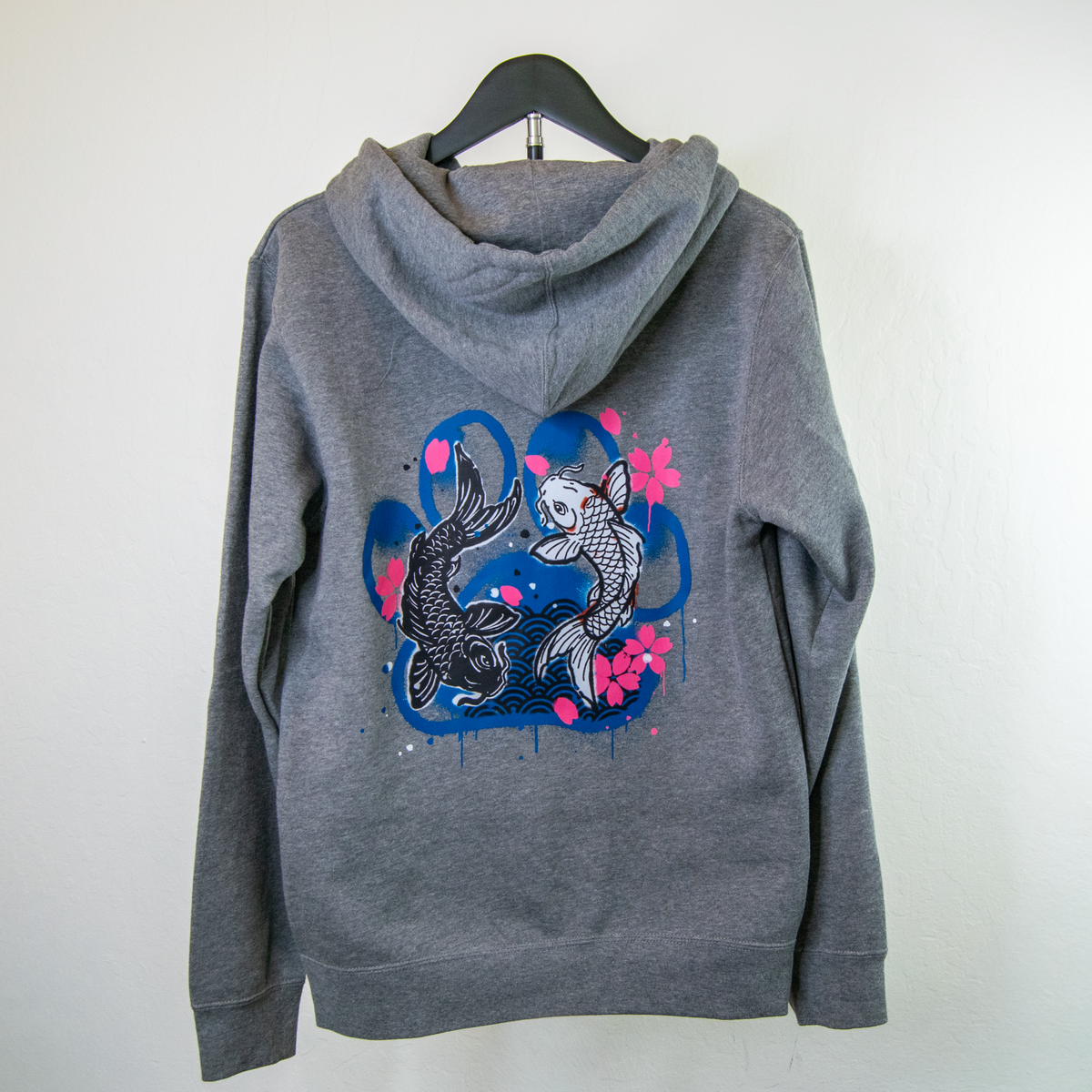 A heather gray pullover hoodie hanging on a black hanger against a plain white wall. The back of the hoodie features a graphic print of two koi fish swimming in blue water with pink cherry blossoms.
