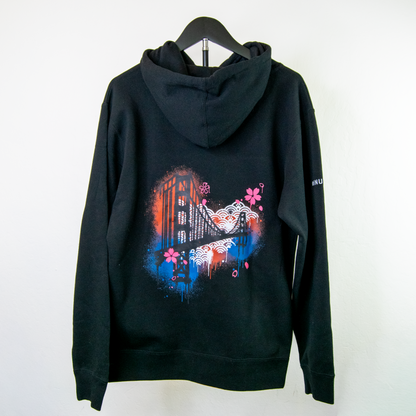 A black pullover hoodie hanging on a black hanger against a plain white wall. The back of the hoodie features a large, colorful graphic print with the golden gate bridge, cherry blossoms, and blue/orange paint splatter details.