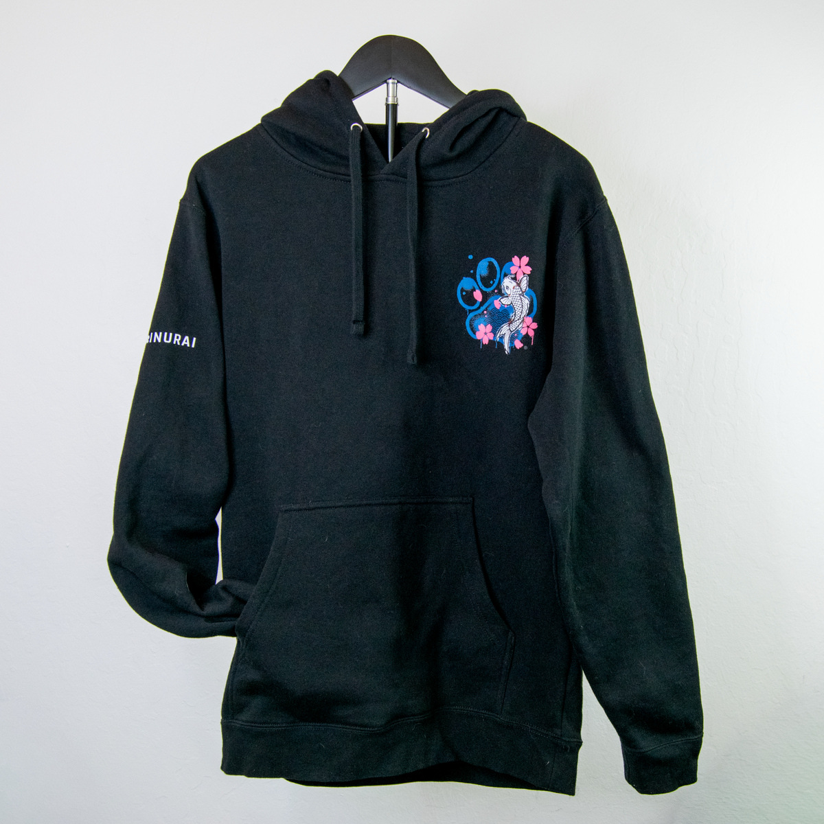 A black pullover hoodie hanging on a black hanger against a plain light gray wall. The hoodie features a colorful graphic on the left chest with blue dog paw, pink cherry blossoms, and a koi fish
