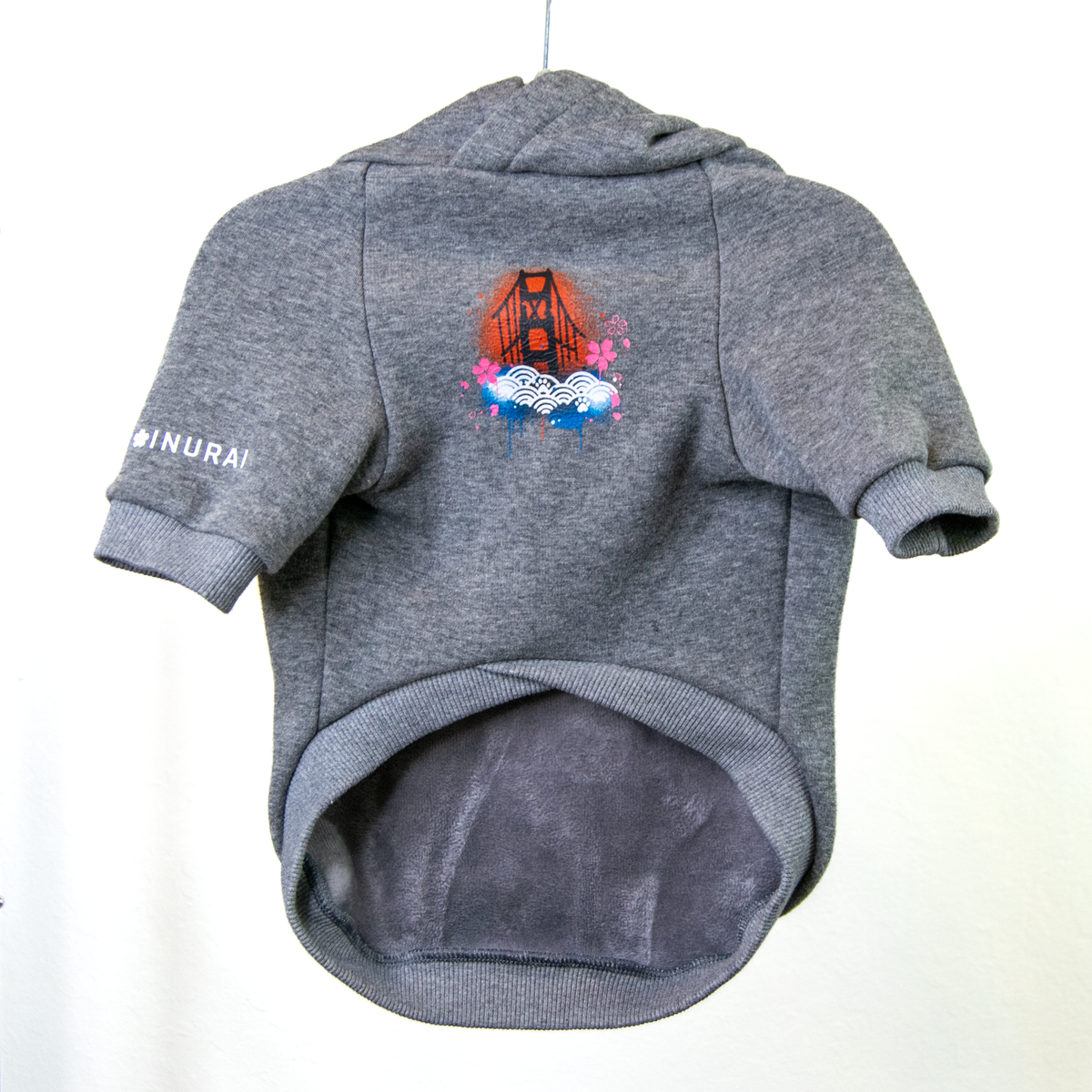 A gray heather pet hoodie with a short-sleeve design, shown hanging against a plain white background. The sweater features a colorful graphic of the Golden Gate Bridge with waves, and the left sleeve has the word 'INURAI' printed in white.