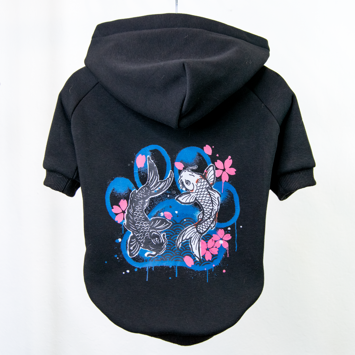 A black pet hoodie shown from the back, featuring a graphic print of two koi fish, pink cherry blossoms, and blue paint splatters.